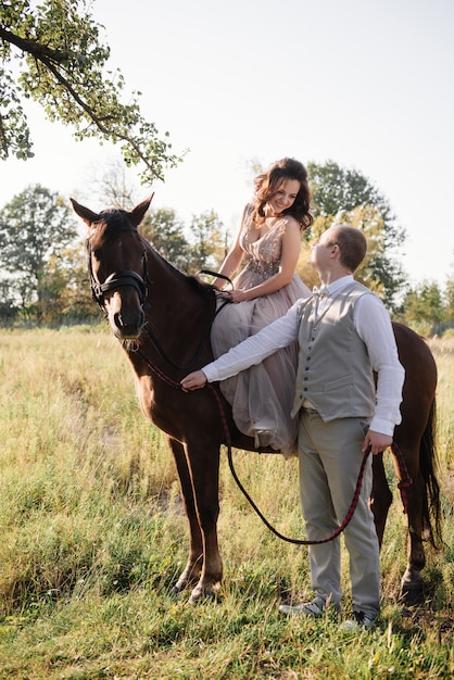 Liefdesverhaal en bruiloft in de buurt van de rivier in een veld bij zonsondergang met bruin paard. De bruid in luchtige jurk is de kleur van stoffige roos. Beige jurk met glitters. bruid en bruidegom omhelzen en kussen.