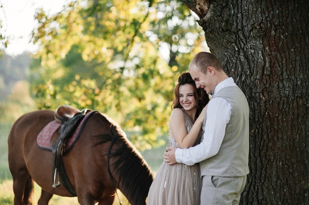 Liefdesverhaal en bruiloft in de buurt van de rivier in een veld bij zonsondergang met bruin paard. De bruid in luchtige jurk is de kleur van stoffige roos. Beige jurk met glitters. bruid en bruidegom omhelzen en kussen.