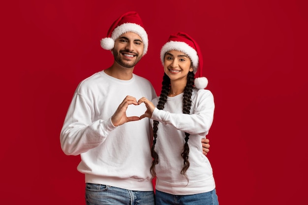 Liefdevol Arabisch koppel in kerstmutsen die een hartsymbool maken met hun handen