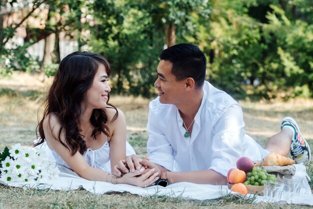 Liefdevol Aziatisch stel dat picknickt in een park, op hun buik ligt en elkaar aankijkt. Man en vrouw rusten in witte kleren in de schaduw van een boom.