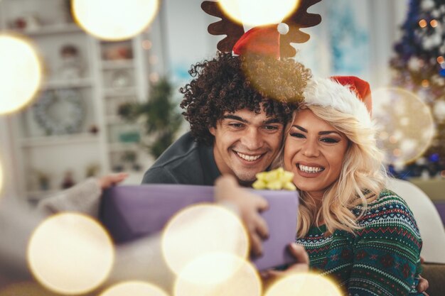 Liefdevol stel gekleed in gezellige warme truien brengt tijd door in de woonkamer op kerst - oudejaarsavond. Jonge man verrast zijn vriendin en geeft haar een geschenkdoos.