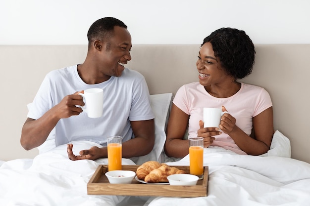 Liefdevolle Afro-Amerikaanse familie man en vrouw ontbijten op bed thuis, kijken naar elkaar en glimlachen. Zwarte minnaars die de ochtend samen in bed doorbrengen, koffie drinken en croissants eten