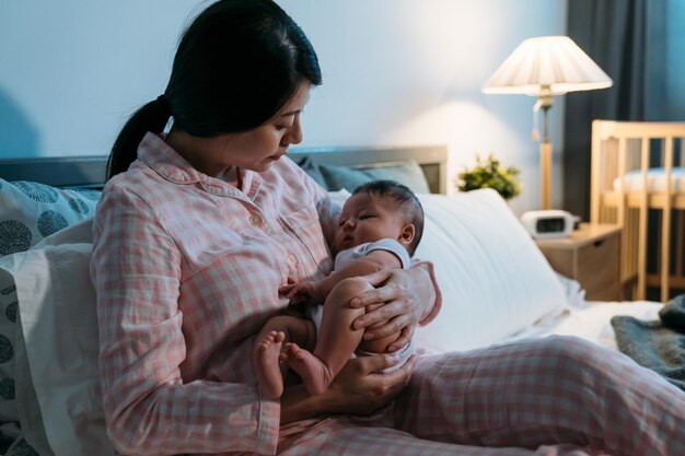 liefdevolle aziatische moeder zittend op bed in pyjama legt haar schattige baby in haar knuffel om te slapen in een gezellige slaapkamer 's nachts thuis.