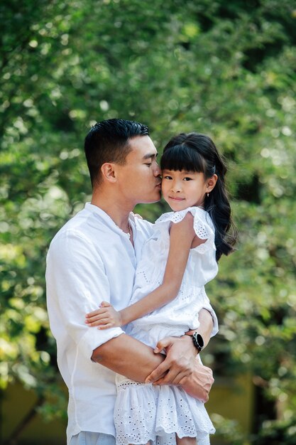 Liefdevolle aziatische vader die zijn dochter in zijn handen houdt en haar op een wang kust. Ze kijkt naar de camera. Ze brengen tijd door in een park, genietend van de laatste warme dagen van de vroege herfst.