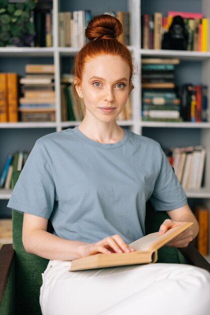 Liefdevolle roodharige jonge vrouw student leest boek geniet van rust op kantoor aan huis, camera kijken. Leuke dame die van boeken geniet bij bibliotheekruimte op achtergrond van boekenplanken.