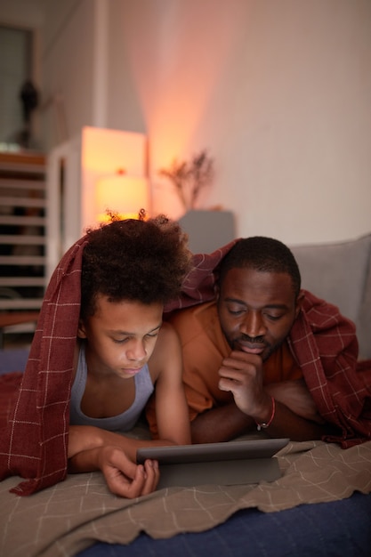 Liefdevolle vader brengt een gezellige avond door met zijn tienerzoon thuis, ontspannend onder de deken, film kijkend online op digitale tablet