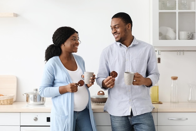 Liefdevolle verwachtende zwarte familie die thee drinkt in de keuken, praat en elkaar aankijkt. vrolijke afro-amerikaanse zwangere vrouw en haar man met mokken, genietend van cacao met koekjes