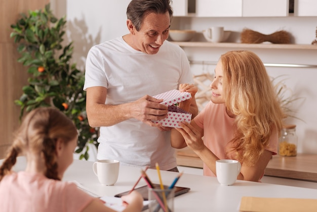 Liefdevolle volwassen vrolijke man genieten van vakantie thuis en cadeau geven aan de vrouw terwijl dochtertje schilderen