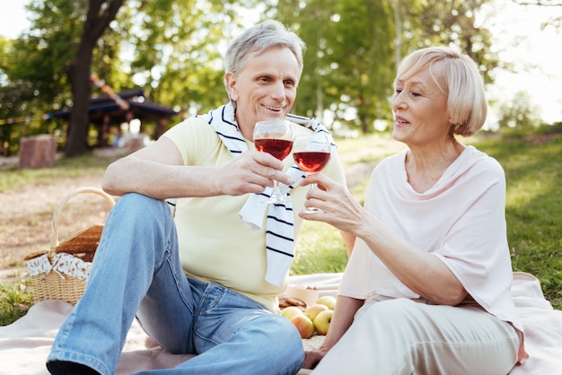 Liefdevolle vrolijke oude paar positiviteit uiten terwijl u geniet van een picknick en het drinken van wijn