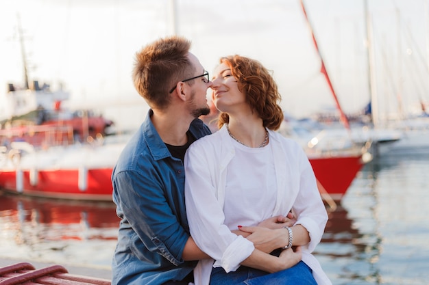 Liefdevolle vrolijke paar liefhebbers kussen zittend op een bankje tegen van jachten club op zee