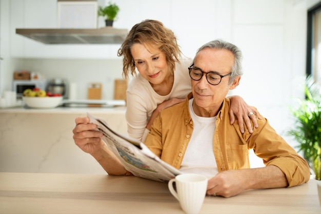 Liefhebbend senior paar dat de ochtend thuis doorbrengt, man die de krant leest, vrouw die echtgenoot omhelst van