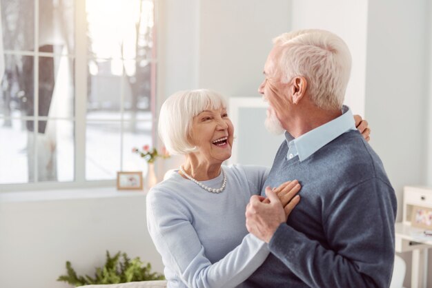 Liefhebbend stel. Vrolijke oudere man en vrouw dansen in de woonkamer terwijl ze breed naar elkaar glimlachen