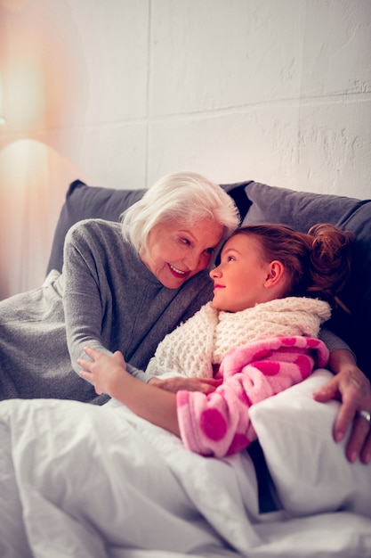 Liefhebbende oma. Zorgzame liefhebbende oma knuffelt haar schattige meisje dat in bed ligt en voelt zich misselijk nadat ze verkouden is