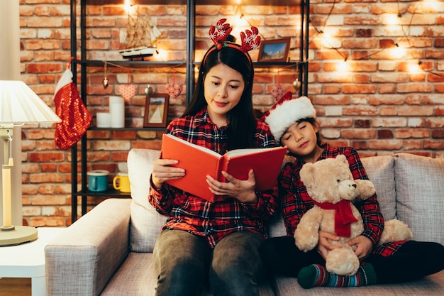 lieve mooie dochter knuffelen speelgoed teddybeer viel in slaap terwijl moeder sprookjesboek las. familie viert verhaaltijd kerstavond in ingericht huis zittend op een comfortabele bank. meisje vertrouw moeder.