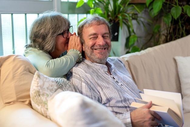 Life of the elderly married couple woman whispering in her\
husband’s ear as he reads a book sitting on the sofa