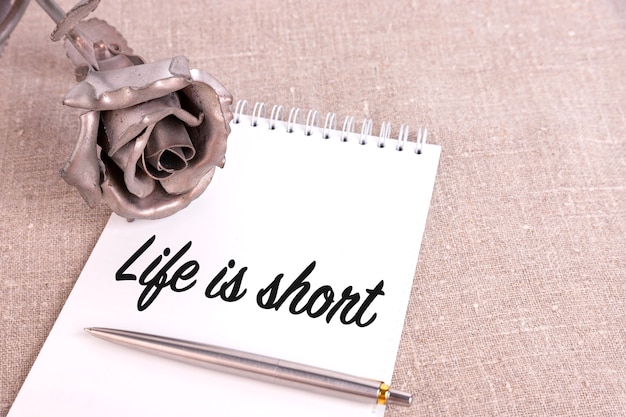 Life is short, the text is written in a notebook lying on a linen linen and an iron rose flower.