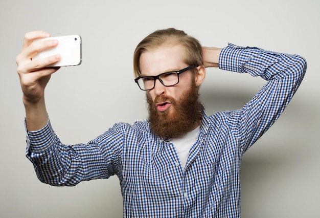 Life style concept: a young man with a beard  in shirt holding mobile phone and making photo of himself while standing against grey background.