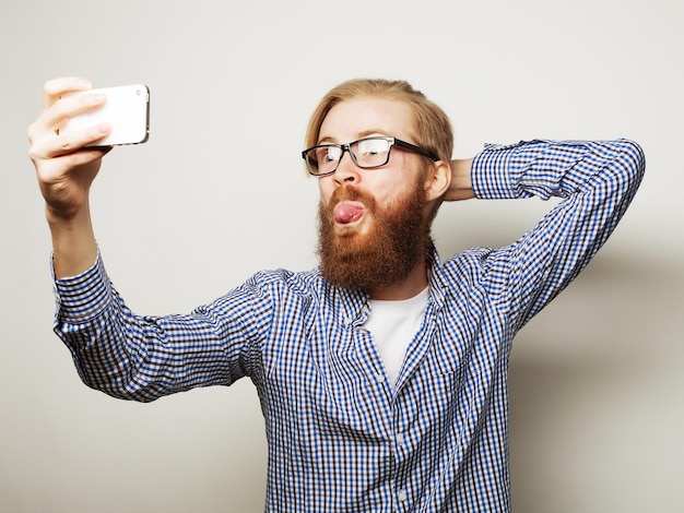 Life style concept a young man with a beard in shirt holding mobile phone and making photo of himself while standing against grey background