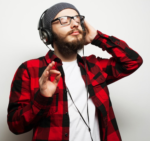 Образ жизни, образование и концепция людей: молодой бородатый мужчина слушает музыку, стоя на сером фоне. Хипстерский стиль.