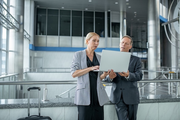 Life style. Serious business good looking woman and man standing near suitcase looking at laptop at airport waiting for departure