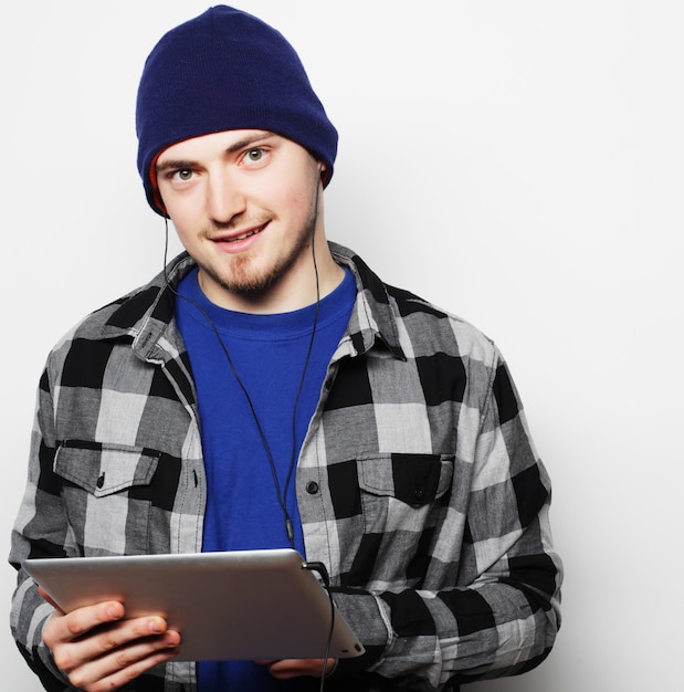 Концепция образа жизни, технологий и людей: красивый молодой человек в рубашке и шляпе работает на цифровом планшете и улыбается, стоя на сером фоне
