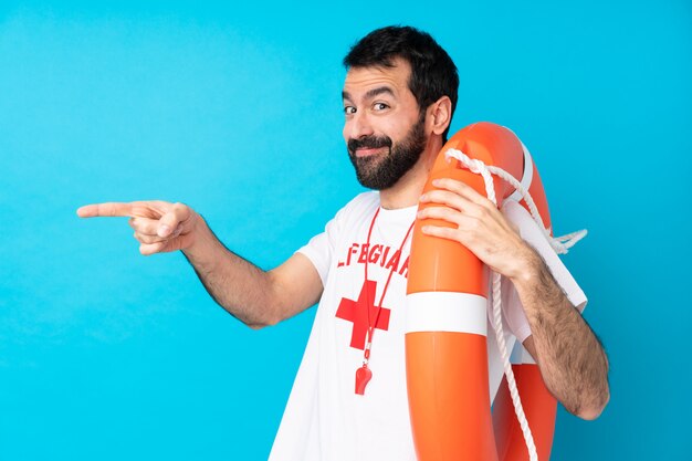 Lifeguard man over isolated blue wall pointing finger to the side and presenting a product