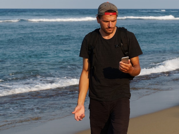 Lifestyle afbeelding. gebruinde man op zoek naar zijn telefoon met een verbaasde uitdrukking op zijn gezicht. wandelen op het tropische strand.