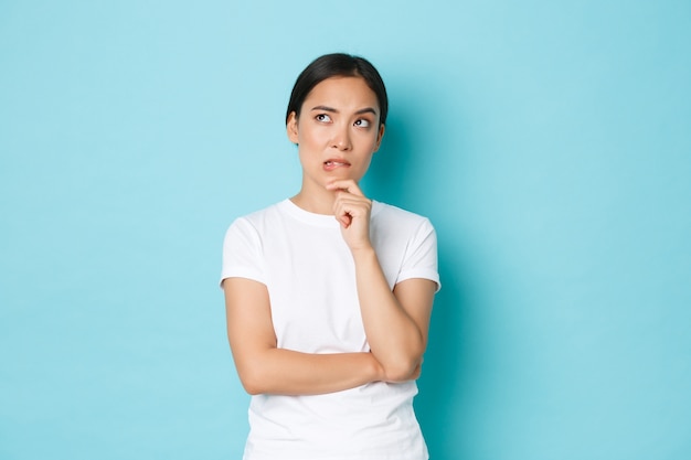 Lifestyle, beauty en shopping concept. Aarzelende en bedachtzame Aziatische vrouw in casual t-shirt op zoek onzeker en nadenkend besluit, staren linkerbovenhoek verbaasd, denkend, blauwe muur.