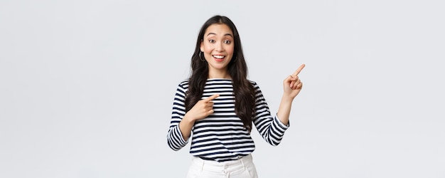 Lifestyle beauty and fashion people emotions concept Surprised happy asian girl standing over white background recommend visit shop pointing fingers right at banner