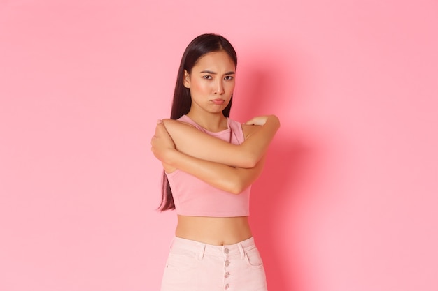 Lifestyle, beauty and women concept. Portrait of defensive and mad cute sulking asian girl ignore someone, unwilling to talk, feeling angry, hugging herself, comforting embrace, pink wall.