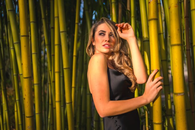 Lifestyle blonde Caucasian girl in a black strappy dress in some beautiful bamboo in the park outdoors the girl perched holding on to the pole and looking at the sun