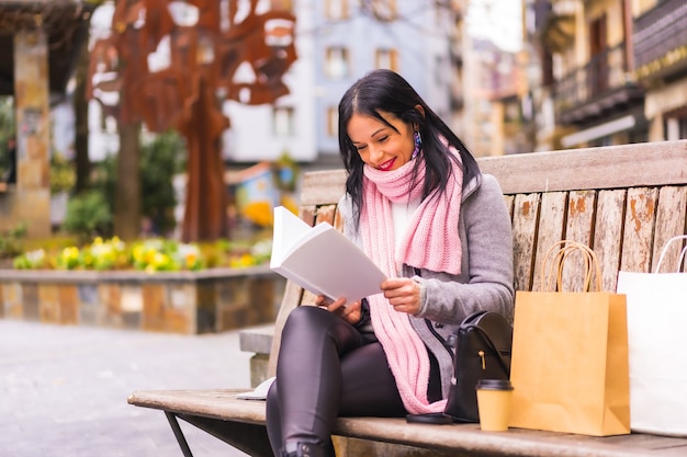 Образ жизни, кавказская девушка брюнетка читает книгу в городском парке, улыбаясь, сидя на скамейке