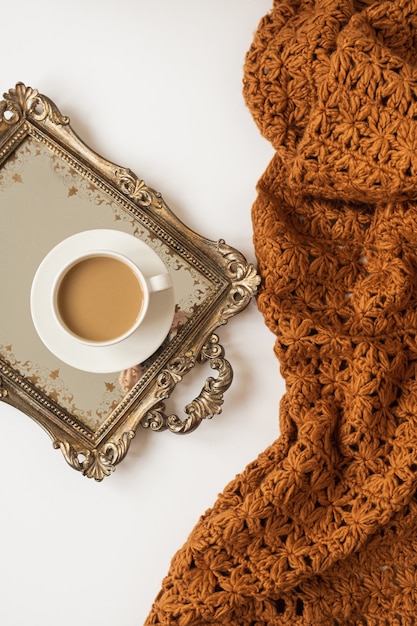 Composizione di stile di vita con una tazza di caffè con latte sul vassoio dorato dell'annata e plaid di coperta marrone lavorato a maglia su priorità bassa bianca.