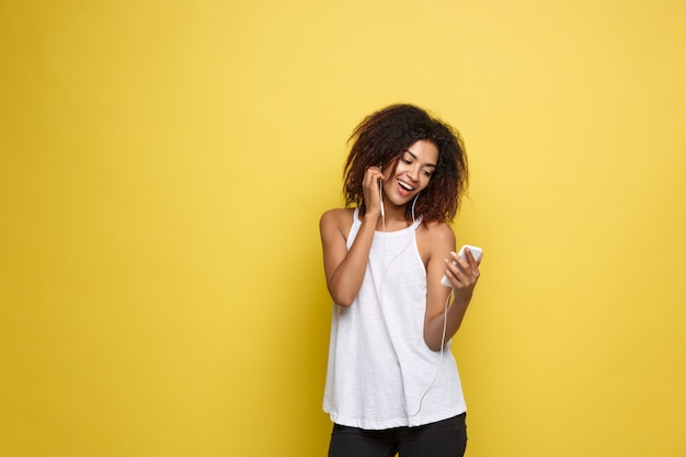 ライフスタイルの概念 - 携帯電話で音楽を聴いて楽しい美しいアフリカ系アメリカ人女性の肖像画。黄色のパステルスタジオの背景。スペースをコピーします。
