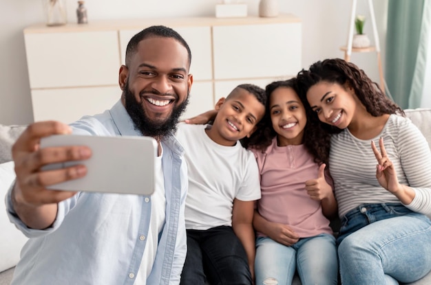 Concetto di stile di vita. ritratto di famiglia nera che prende selfie insieme. genitori allegri che posano con i loro figli e sorridono alla telecamera, seduti sul divano nel soggiorno di casa, papà che tiene il cellulare