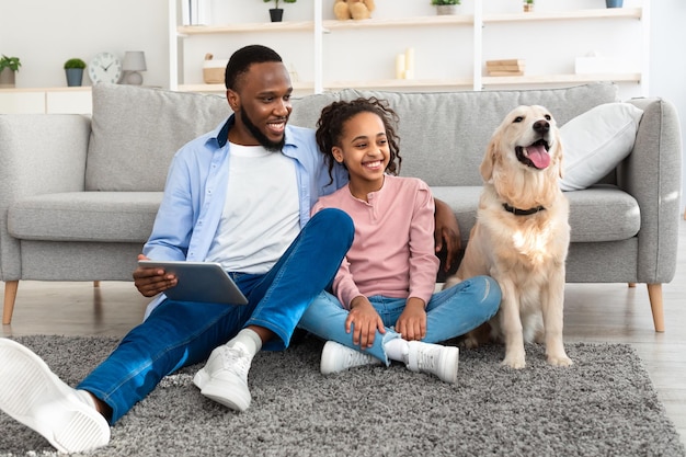 Lifestyle Concept. Portrait of smiling black man using digital tablet, sitting on the floor carpet and embracing daughter, looking at happy dog. Family spending time at home, having fun