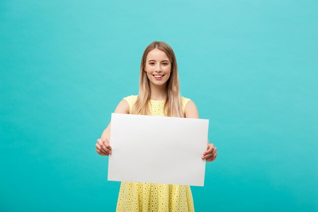 Lifestyle concept young beautiful girl smiling and holding a blank sheet of paper dressed in yellow isolated on pastel blue background person