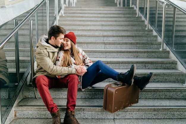 Lifestyle couple of happy romantic hipsters in love. Young pair of lovers with emotional faces. Man with girlfriend, suitcases wait for train on stairs.