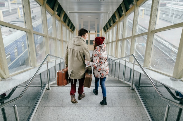 Lifestyle couple of young happy lovers carry vintage brown suitcases at railway station. Pair of stylish cheerful hipsters going down the stairs.