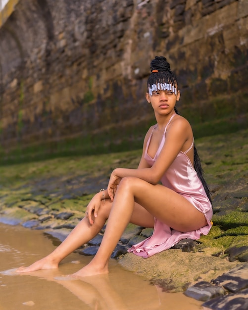 Photo lifestyle of a dominican ethnic girl with braids with a beautiful pink dress. fashionable posing enjoying summer on the beach sitting on the sand