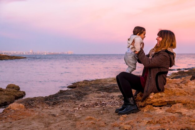 Stile di vita di una famiglia sulla spiaggia un bambino che si diverte con sua madre seduta in riva al mare al tramonto Foto Premium