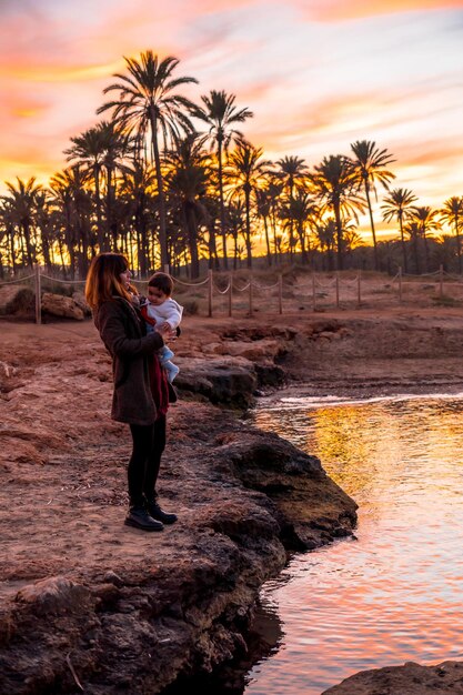 Stile di vita di una famiglia sulla spiaggia giovane madre con il suo bambino in riva al mare al tramonto sulla spiaggia di palme