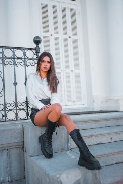 Lifestyle, fashionable pose of a young brunette in a black leather skirt and shirt sitting on some stairs with seductive gaze