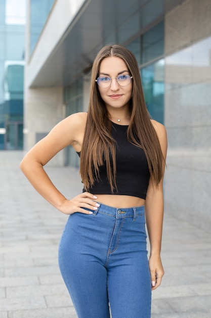 Lifestyle, femininity, body and people concept - A pretty, smiling, curvy girl in glasses, black tank top, and blue jeans standing on the street with one hand around her waist