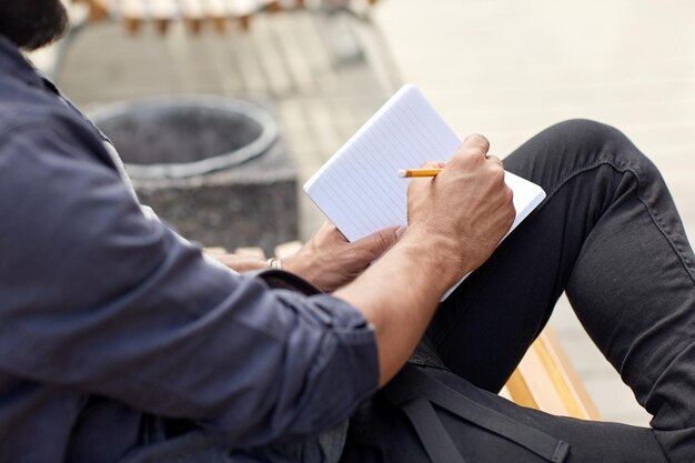 lifestyle, freelance, inspiration and people concept - close up of man with bag writing to notebook or diary sitting on city street bench