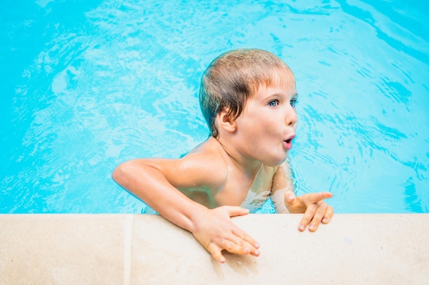 Lifestyle funny portrait boy swim in pool hold edge bright blue water surprised excited look point finger childhood behavior aqua park entertainment summer camp training leisure activity