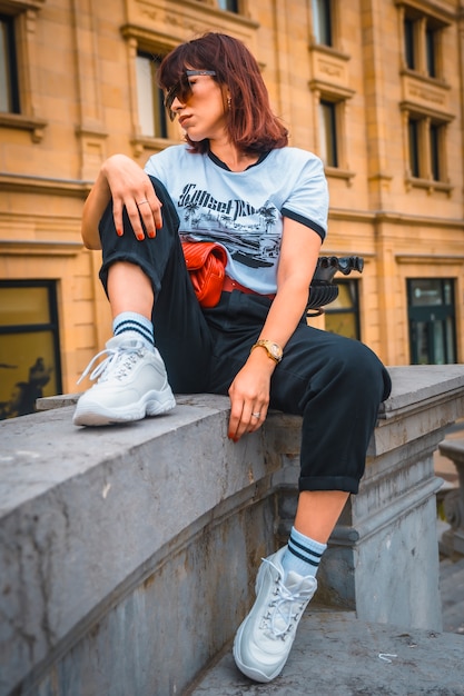 Lifestyle in de stad. Een jonge brunette in zonnebril, een wit T-shirt, een rode heuptas, een zwarte broek en witte sneakers. Modieuze baars zittend op een loft