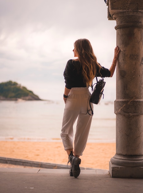 Lifestyle in de stad met een blond meisje in een witte broek en een leren jas vlakbij het strand. foto's naast een kolom die naar de zee kijkt en naar links