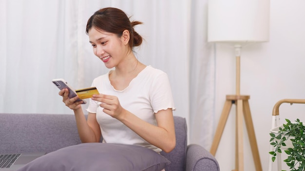 Lifestyle in living room concept Asian woman holding credit card and use phone to online shopping