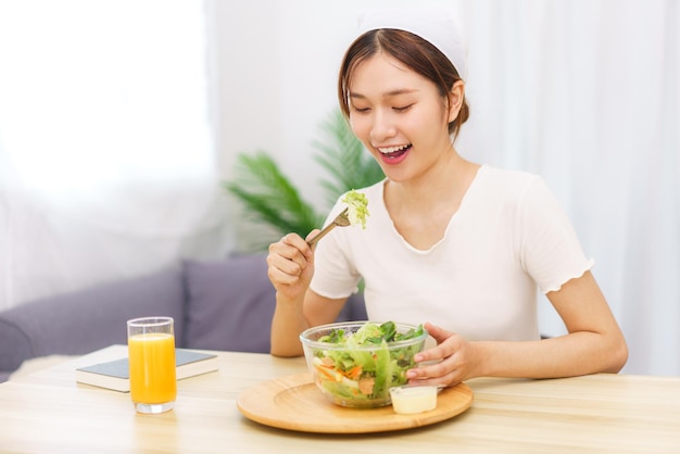 リビングルームのコンセプトのライフスタイル野菜サラダとオレンジジュースを食べる若いアジアの女性